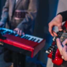 Close-up of hands playing electronic keyboard during live music performance with blurred bassist in background under blue lighting.